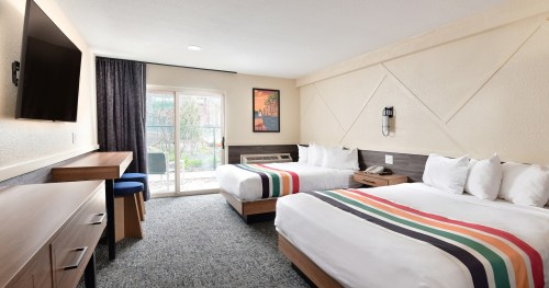 A cozy hotel room with two beds, a TV, desk, and sliding door leading to an outdoor area. Bright and inviting decor.