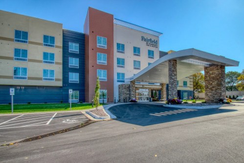 Modern hotel exterior with a stone entrance, large windows, and a clear blue sky. Parking area in front.