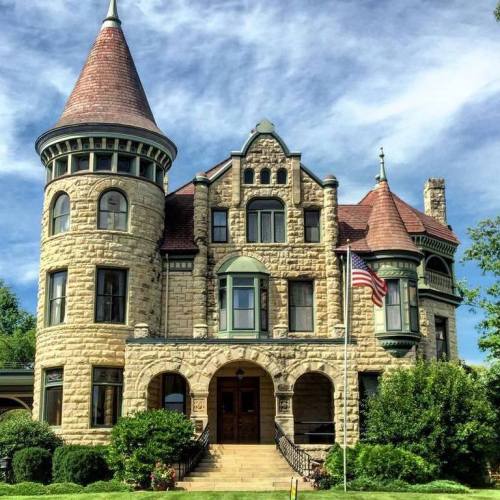 A grand stone mansion with a turret, red roof, and American flag, surrounded by lush greenery and a blue sky.