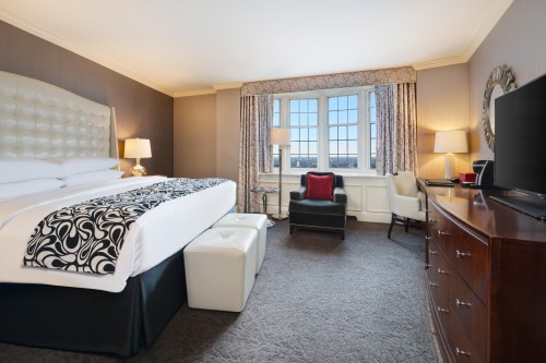 A stylish hotel room featuring a large bed, armchair, window with curtains, and a wooden dresser.