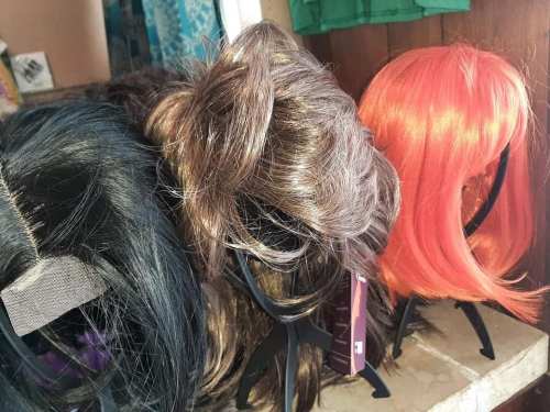 Three colorful wigs displayed on stands: black, brown, and bright orange, arranged neatly on a surface.
