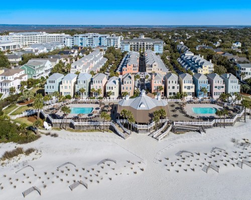 Aerial view of colorful beachside homes with pools, surrounded by sandy beach and palm trees under a clear blue sky.