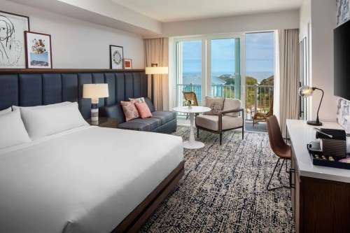 A modern hotel room featuring a king bed, seating area, and balcony with ocean views. Bright and inviting decor.