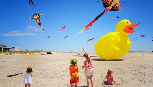 Children watch colorful kites flying in the sky, including a large yellow duck, on a sunny beach day.