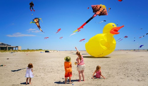 Children watch colorful kites flying in the blue sky at the beach, including a large yellow duck kite.