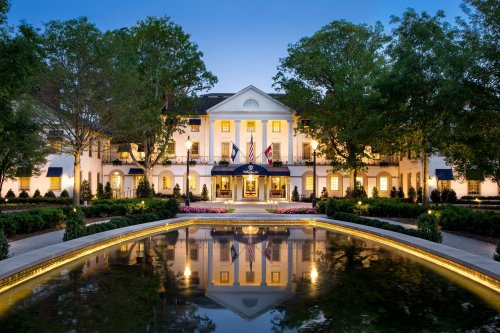 A grand white building with columns, surrounded by trees and a reflecting pond, illuminated at dusk.