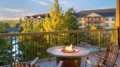 A cozy balcony with a fire pit overlooks a pool and lush trees, with buildings in the background under a sunset sky.