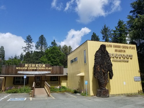 A yellow building with a wooden sign reading "Museum" and a Bigfoot statue outside, surrounded by trees and blue sky.