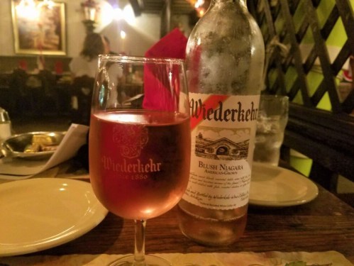 A glass of Blush Niagara wine on a table with a bottle of Wiederkehr wine in the background, set in a cozy restaurant.