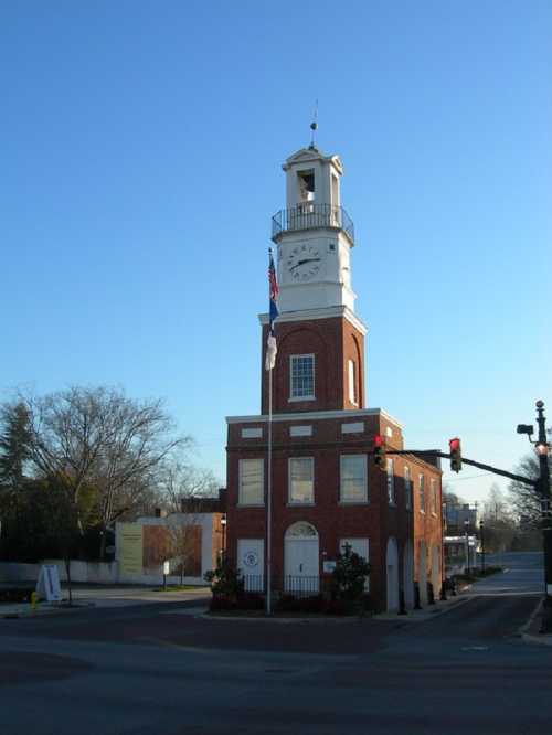 Historic brick clock tower with a weather vane, surrounded by trees and a clear blue sky. Traffic light visible nearby.