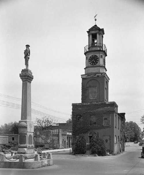 Historic clock tower with a statue nearby, surrounded by buildings and trees in a small town setting. Black and white photo.