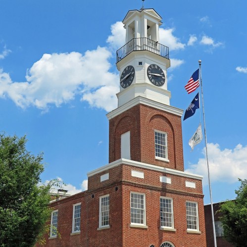Historic clock tower with a flag, surrounded by trees and a blue sky with clouds.