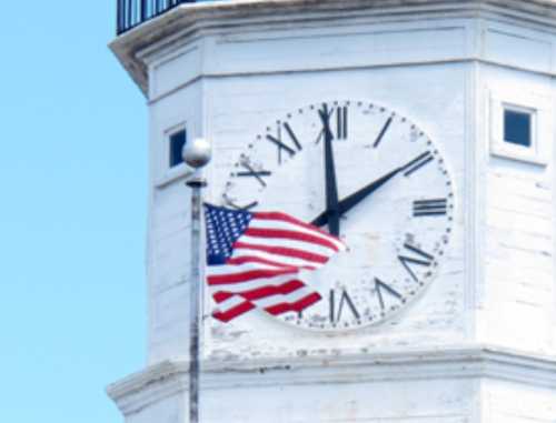 A close-up of a clock tower with a large clock face and an American flag waving in front against a clear blue sky.