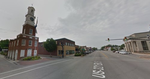 A street view featuring a clock tower, shops, and traffic lights in a small town setting under a cloudy sky.