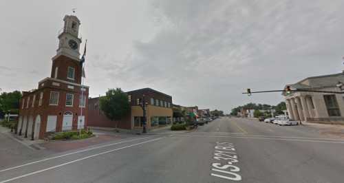 A street view featuring a clock tower, shops, and traffic lights in a small town setting under a cloudy sky.