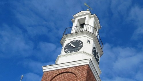 A clock tower with a weather vane against a blue sky, featuring a large clock face and brick base.