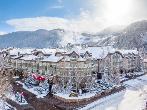 A snowy mountain resort with charming architecture, surrounded by trees and hills under a bright blue sky.