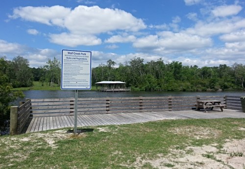 A wooden dock at Wolf Creek Park with a sign, overlooking a calm waterway and surrounded by greenery under a blue sky.