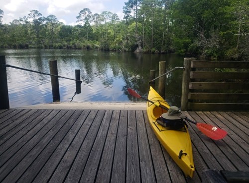 A yellow kayak rests on a wooden dock by a calm, reflective river surrounded by lush greenery.
