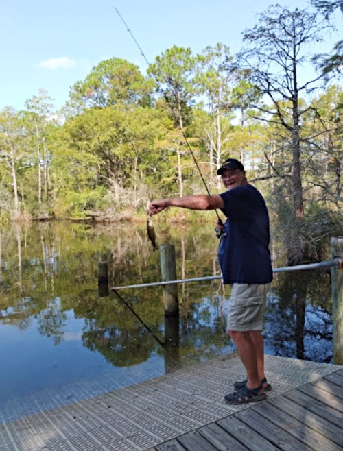A man stands on a dock, holding a fish he caught, with trees and water in the background.