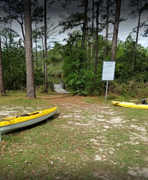 Two yellow kayaks are parked on a grassy area near a wooded path leading to a waterway and a sign.