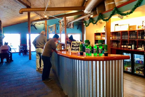A rustic bar with patrons enjoying drinks, surrounded by shelves of beverages and festive decorations.