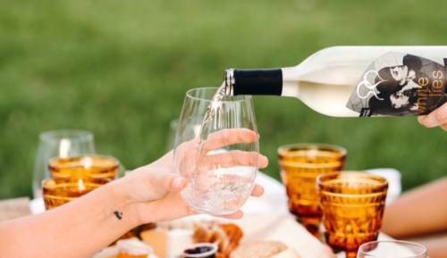 A hand pours white wine into a glass at a picnic, surrounded by amber glasses and a spread of food on a grassy surface.