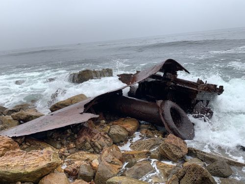 A rusted shipwreck partially submerged on rocky shore, with waves crashing against it under a foggy sky.