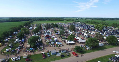 Aerial view of a bustling outdoor market with numerous tents, cars, and people amidst green fields and trees.