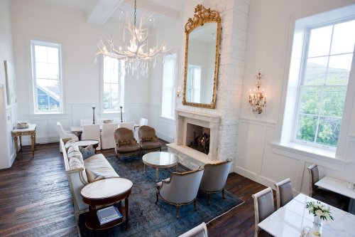 Bright, elegant living room with a chandelier, large mirror, and cozy seating around a coffee table. Large windows let in light.