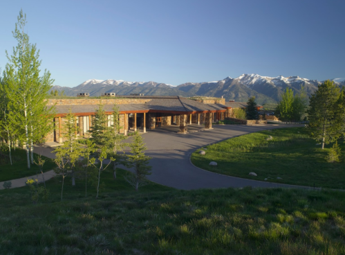 A scenic view of a modern building surrounded by trees, with snow-capped mountains in the background under a clear sky.