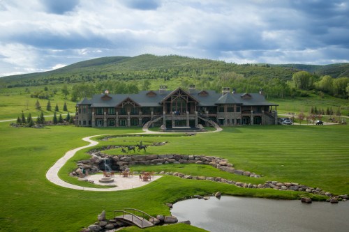 A large, elegant lodge surrounded by green lawns, a pond, and mountains under a cloudy sky.