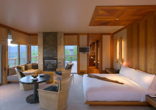 A modern hotel room featuring a king bed, seating area, fireplace, and large windows with a scenic view.