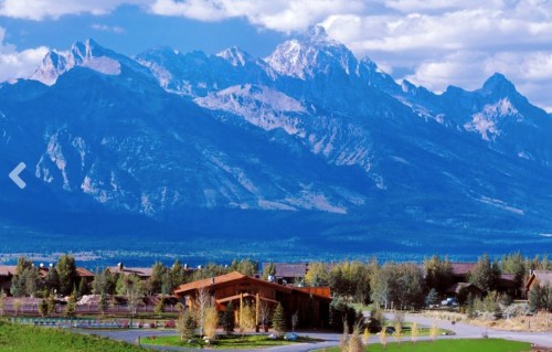 A scenic view of mountains with a wooden building in the foreground, surrounded by trees and a clear blue sky.