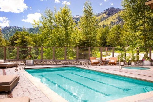 A serene outdoor pool surrounded by trees and mountains, with lounge chairs and a wooden table nearby.