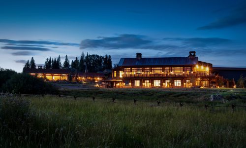 A rustic lodge illuminated at dusk, surrounded by trees and a grassy field under a twilight sky.