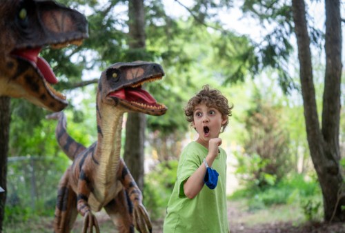 A surprised child in a green shirt stands between two dinosaur costumes in a forested area.