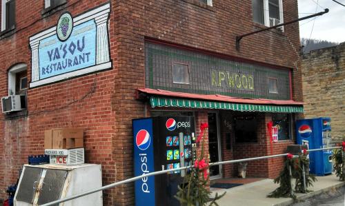 Exterior of Yasou Restaurant with a brick facade, green awning, and Pepsi vending machines outside, decorated for the holidays.
