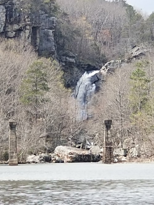 A waterfall cascades down rocky cliffs, surrounded by bare trees, with a calm lake in the foreground.