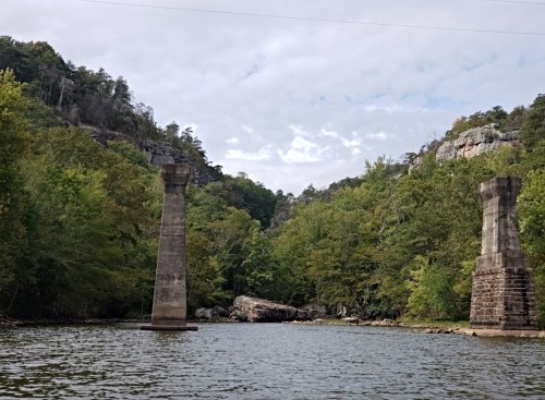Two tall, weathered stone pillars stand in a calm river, surrounded by lush green trees and rocky cliffs under a cloudy sky.