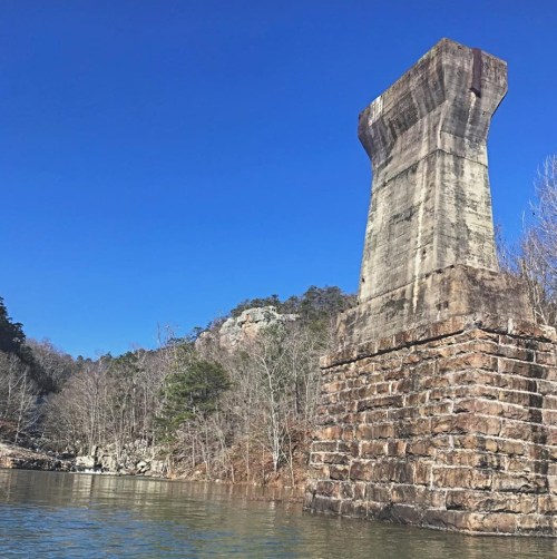 A tall, weathered stone pillar stands by a calm river, surrounded by trees and rocky hills under a clear blue sky.