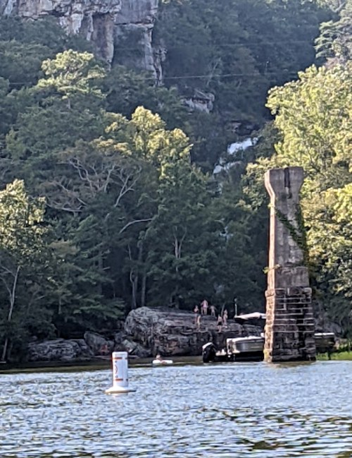 A serene lake scene with a rocky shore, a tall stone pillar, and people enjoying the outdoors near a waterfall.