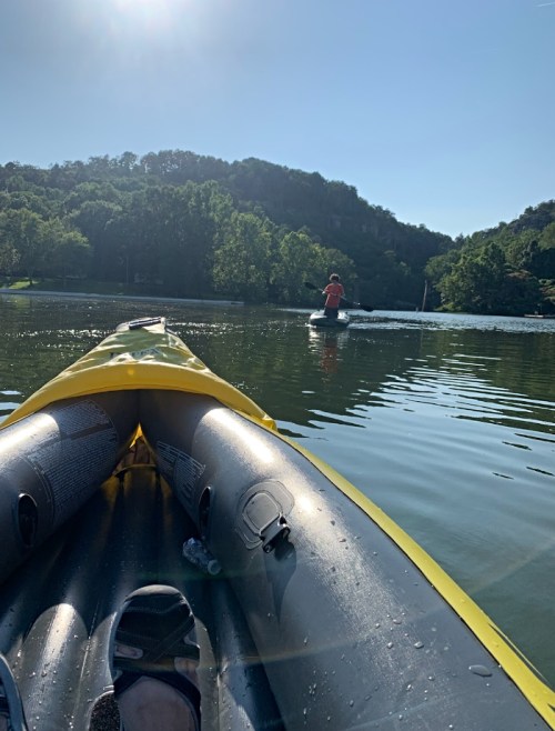 A view from a yellow kayak on calm water, with a person in the distance paddling on a sunny day.
