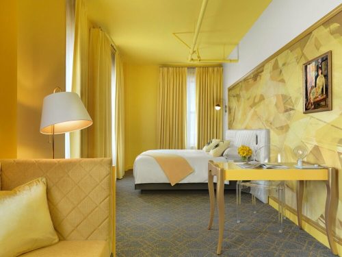 Bright yellow hotel room featuring a bed, desk, and cozy seating area with large windows and modern decor.