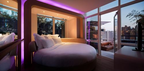 Modern hotel room with a round bed, purple lighting, large windows, and a balcony overlooking the city at sunset.