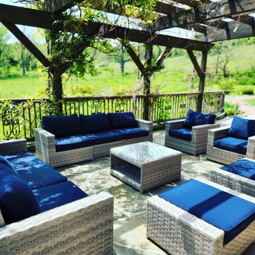 A cozy outdoor seating area with wicker furniture and blue cushions, surrounded by greenery and a wooden pergola.