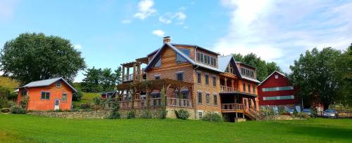 A large, multi-story house with a wooden deck, surrounded by green grass and trees, with a red barn in the background.