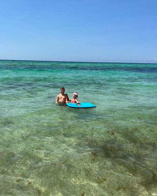 Two children play in shallow, clear water, one holding a blue boogie board, with a sunny sky and ocean in the background.