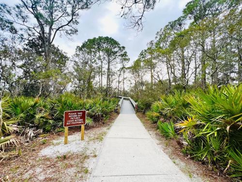 A paved path leads through lush greenery to a wooden boardwalk, with a sign asking visitors to stay on designated paths.