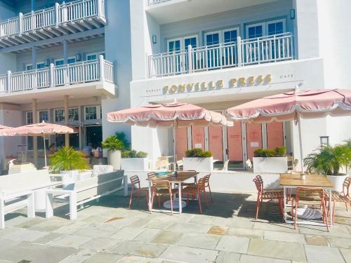 Outdoor seating area with pink umbrellas in front of a café named "Bonville Press," featuring white furniture and potted plants.